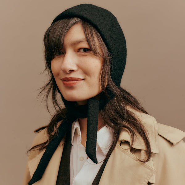 Woman wearing a black bonnet and beige coat against a brown background