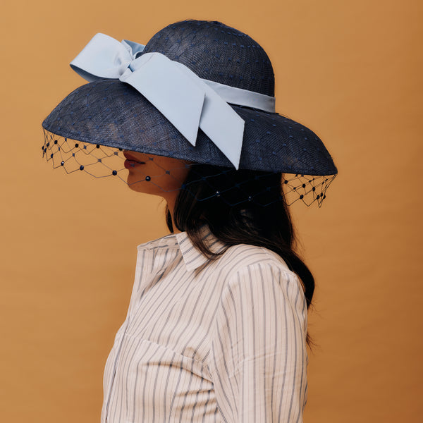 Woman wearing a navy blue hat with a large light blue bow against a brown background