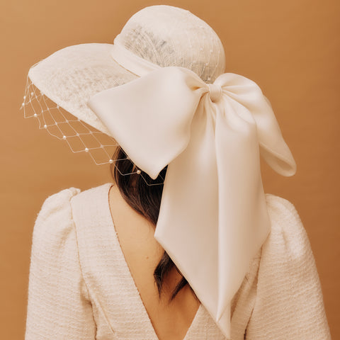 Woman wearing a large cream-colored hat with a bow and veil against a brown background