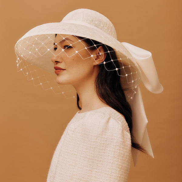 Woman wearing a white wide-brimmed hat with a veil against a brown background
