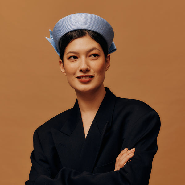 Woman wearing a black outfit and light blue pill box hat against a brown background