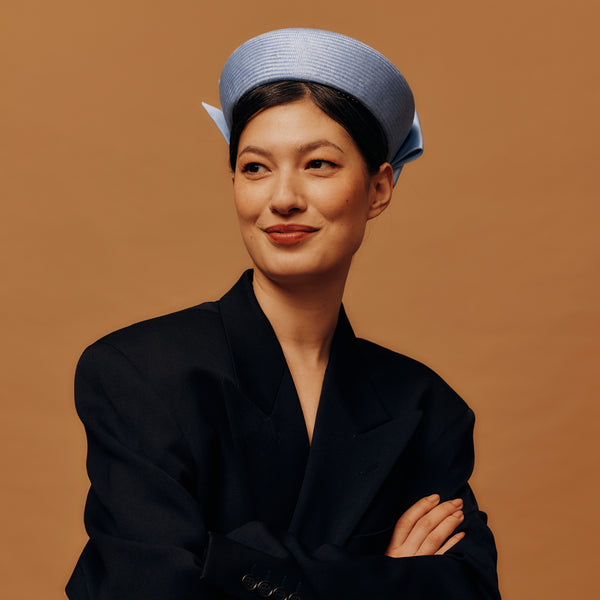 Woman wearing a black coat and blue pill box hat against a brown background