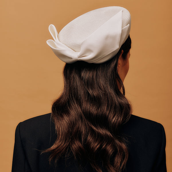 Person wearing a white pillbox hat with a bow on a beige background