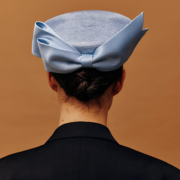 Person wearing a blue straw pill box hat with a large bow on a brown background