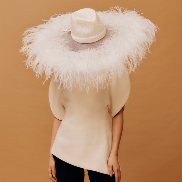 Person wearing a beige outfit with a feathered white hat against a brown background