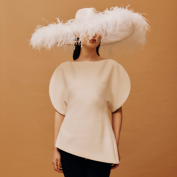Person wearing a large white feathered hat and a beige top against a brown background