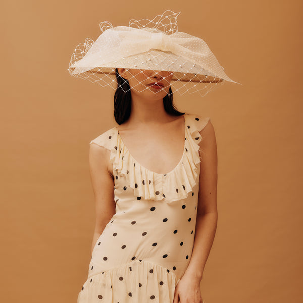Woman wearing a white dress with black polka dots and a large white hat against a brown background