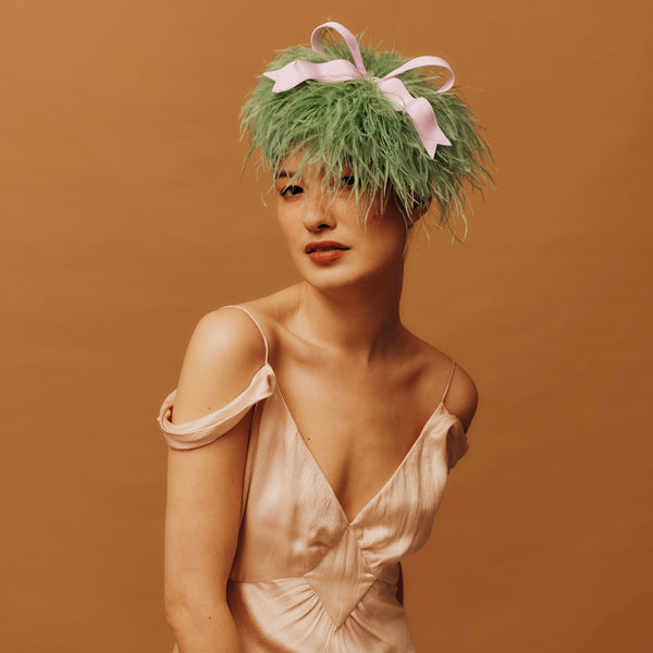 Woman wearing a green feathered headpiece with a pink bow against a brown background