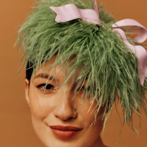 Person wearing a green feathered headpiece with a pink bow on a brown background