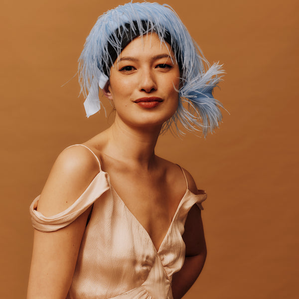 Woman with blue feather headband wearing a beige dress against a brown background